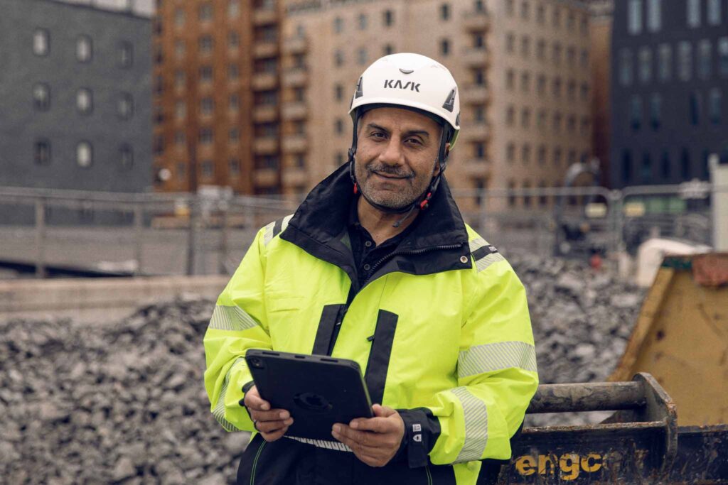 Byggledare med hjälm och reflexjacka står på byggarbetsplats och använder surfplatta för digital dokumentation