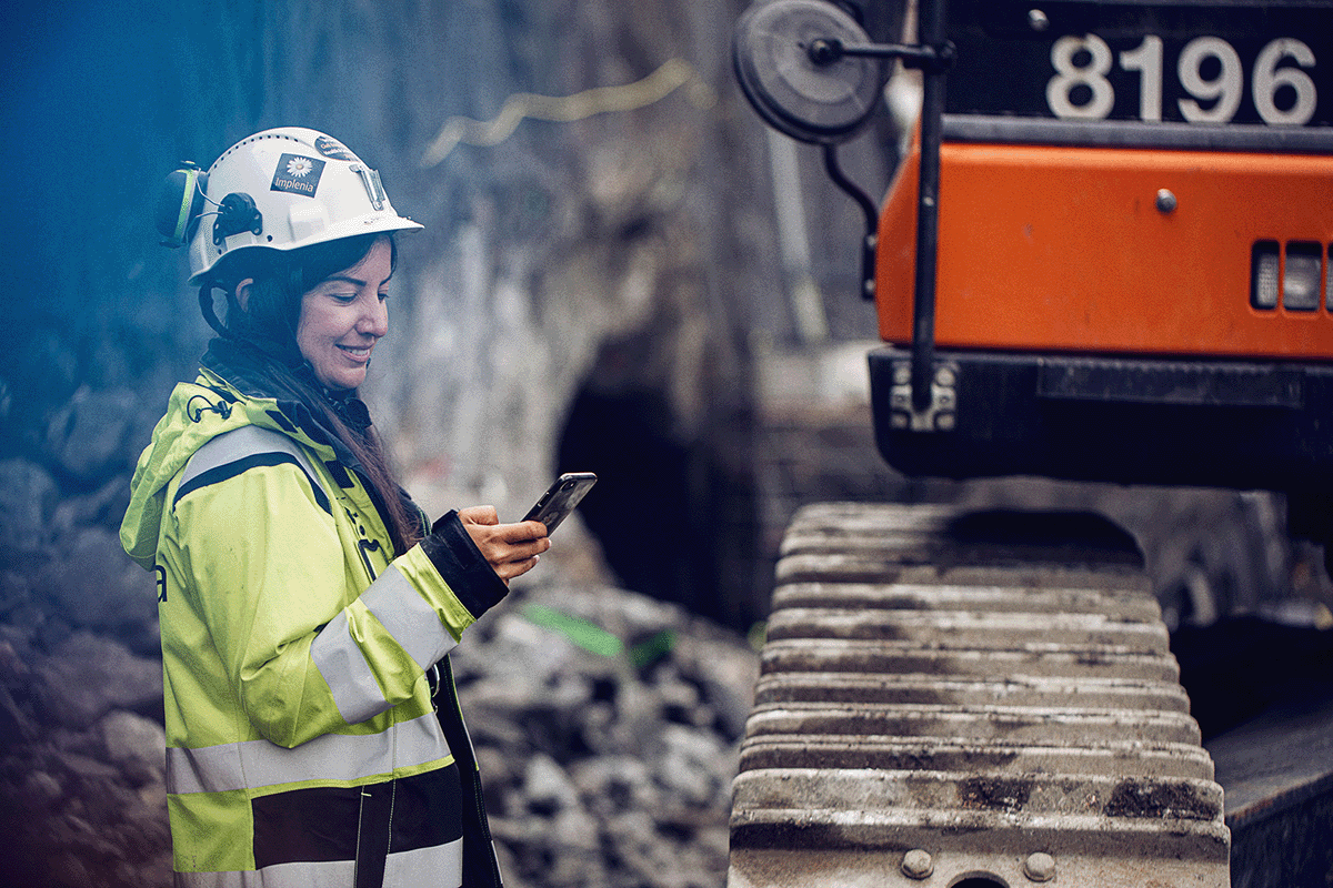 Kvinna i byggarbetskläder framför grävmaskin kollar Infobric Workorder i sin mobil